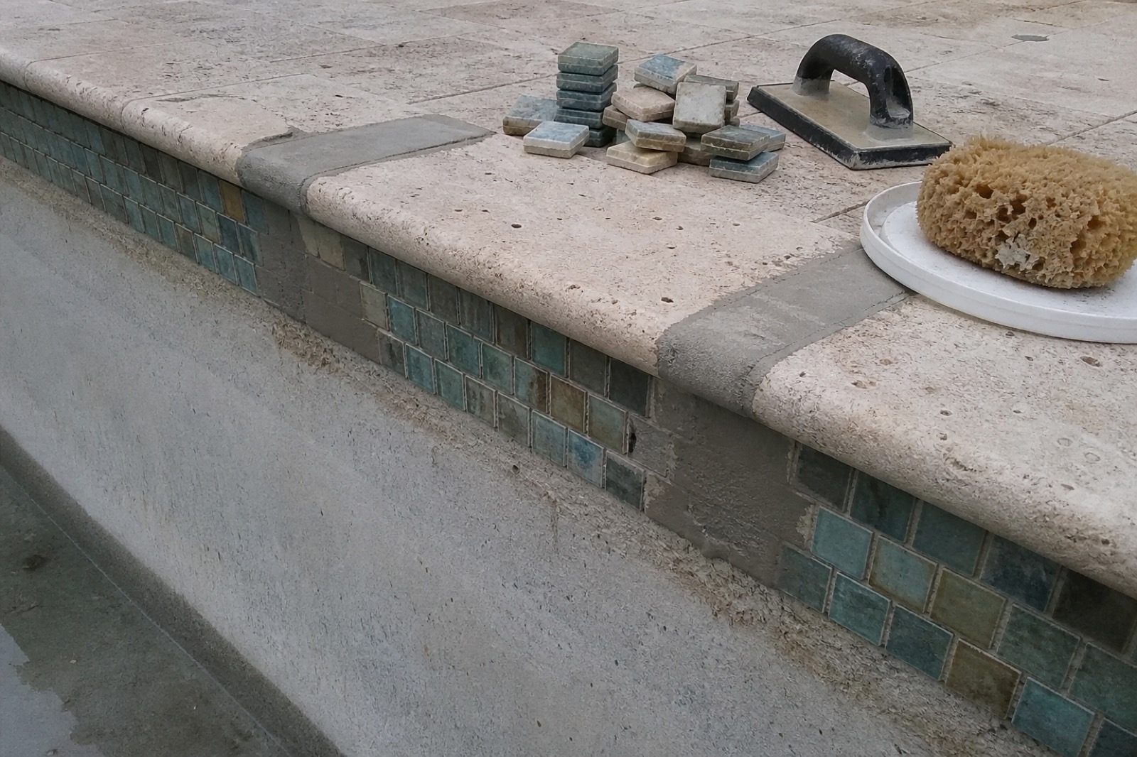 Waterline tile and travertine coping mid-repair, with replacement tile, grout float, and sponge on the deck